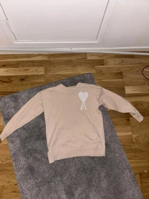 Beige stickad tröja från Ami - Säljer en beige stickad tröja från Ami med deras ikoniska hjärta och A-logga broderad på bröstet. Tröjan har rund halsringning, långa ärmar och ribbade muddar. Perfekt för dig som gillar stilrena och trendiga plagg. Passar även i xs väldigt bra!