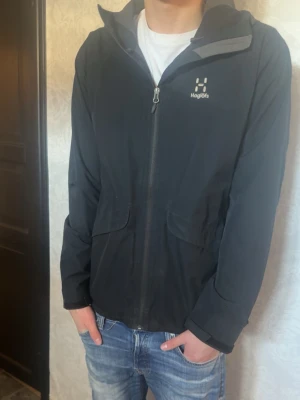 Svart vindjacka från Haglöfs - Säljer en svart vindjacka från Haglöfs med hel dragkedja och två stora fickor framtill. Jackan har hög krage och diskret logga på bröstet. Materialet är lätt och smidigt, perfekt för att skydda mot vind. Passar dig som gillar en enkel och stilren look.