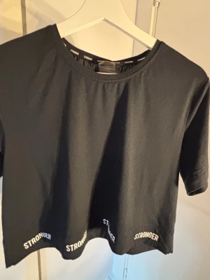 Svart crop top från Stronger - Säljer en svart crop top från Stronger med rund halsringning och korta ärmar. Nedtill finns vita Stronger-loggor tryckta runt hela fållen. Materialet är mjukt och stretchigt, perfekt för träning eller chill. Enkel och stilren design som funkar till det mesta.