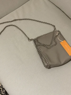 Grå/beige axelväska med kedja Gina Tricot young - Snygg grå axelväska från Gina Tricot Young med silverfärgad kedjerem och dekorativa kedjedetaljer längs kanterna. Väskan är i ett mjukt, skinnliknande material och har en stilren, fyrkantig form. 