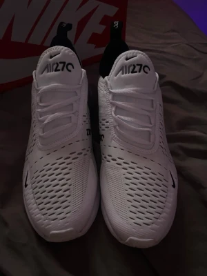 Nike Air Max 270 vita sneakers - Nike Air Max 270 sneakers i vitt med svart logga och detaljer. Ovandelen är i mesh för bra ventilation och sulan har den ikoniska Air-enheten för extra komfort. Snygg design med dragflik på hälen och klassisk snörning. Perfekta för dig som gillar streetwear och sportig stil.