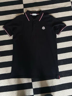 Svart Moncler pikétröja med logga - Snygg svart pikétröja från Moncler med klassisk krage och knäppning framtill. Kragen har vita och röda ränder och bröstet pryds av Monclers ikoniska logga. Perfekt för en clean och sportig look.