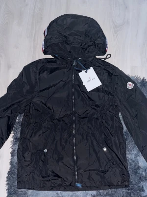 Svart vindjacka från Moncler - Snygg svart vindjacka från Moncler med huva och dragkedja framtill. Jackan har Moncler-logga på ärmen, två fickor med tryckknappar och en vit-röd-blå rand på huvan. Tillverkad i lätt, glansigt material som står emot vind.