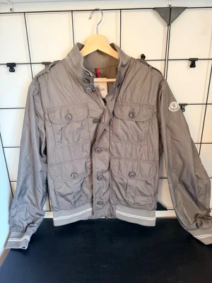 Moncler Field Jacket grå - Snygg och stilren grå Moncler jacka i lätt, glansigt material med exklusiv känsla. Fyra rymliga fickor framtill, knappstängning samt ribbade muddar för bra passform. Moncler-logga på ärmen som bekräftar äkthet. Perfekt för vår/sommar och passar både streetwear och en mer uppklädd stil.  ✔ 100% äkta ✔ Lätt och bekväm ✔ Fyra praktiska fickor ✔ Stilren premiumdesign
