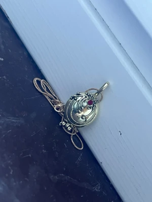 Elena gilbert necklace Guldfärgad - Säljer ett guldfärgat medaljonghalsband med en oval form och dekorativa detaljer på framsidan. Medaljongen har en liten röd sten och kan öppnas för att lägga in ett foto eller minne. Kedjan är tunn och elegant, perfekt för att ge din stil en personlig touch.