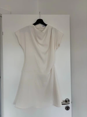 Vit kortklänning från Zara - Stilren vit kortklänning från Zara med draperad halsringning och lös passform. Klänningen har korta ärmar och är tillverkad i ett mjukt, lätt tyg som ger ett elegant fall. Perfekt för dig som gillar minimalistisk och modern stil. Klänningen är använd en gång och är därför i ett super skick💗💗