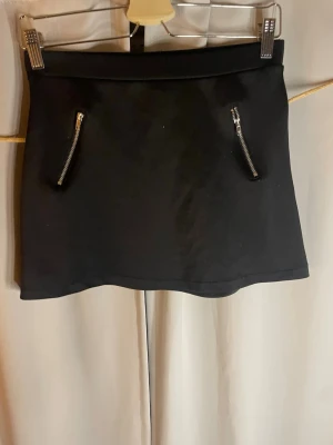 Svart kortkjol med dragkedjor - Stilren svart kortkjol med två sneda dragkedjor framtill som ger en cool detalj. Kjolen har en rak passform och bred linning. Perfekt att matcha med en crop top eller oversized hoodie.