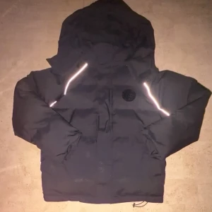 Svart pufferjacka - Svart pufferjacka med stor huva och reflekterande detaljer vid dragkedjorna. Jackan har en rund logotyp på bröstet och är vadderad för extra värme. Perfekt för kalla dagar och har en modern, boxig passform.