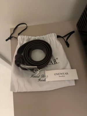 Mörkbrunt skärp från ONEWear - Snyggt mörkbrunt skärp från ONEWear Sweden i klassisk design. Skärpet är tillverkat i skinn och mocka. Den har ett stilrent silverfärgat spänne. Perfekt accessoar för att lyfta din outfit och funkar till både jeans och kostymbyxor.
