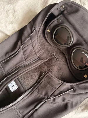 Grå vindjacka med goggles från C.P. Company - Säljer en grå vindjacka från C.P. Company med ikoniska goggles i huvan. Jackan har dragkedja framtill, justerbar huva med snörning och mjukt fleecefoder på insidan. Perfekt för dig som vill ha en unik och funktionell jacka. SITTER SOM M/L