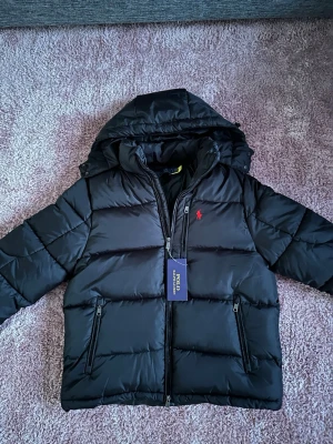 Svart dunjacka från Polo Ralph Lauren - Snygg svart dunjacka från Polo Ralph Lauren med huva och två dragkedjeförsedda fickor. Jackan har det klassiska röda Polo-logotypen broderad på bröstet och en quiltad design som ger en modern look. Perfekt för kalla vinterdagar.