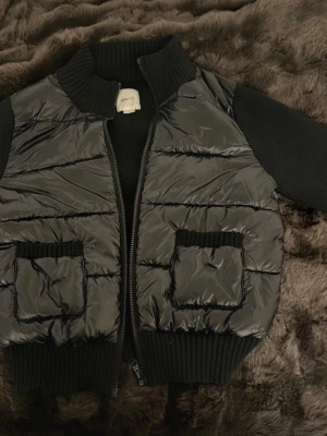 Svart stickad pufferjacka från Gina Tricot - Snygg svart stickad pufferjacka från Gina Tricot med stickade muddar vid krage, ärmslut och nederkant. Jackan har två stora fickor framtill med ribbade detaljer och dragkedja. Materialet är glansigt och quiltat, perfekt för kalla dagar. Storlek L i Young men S/M i dam storlek