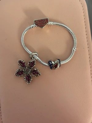 Stranger Things armband med berlock - Snyggt armband i silverfärg med Stranger Things-logga och en stor blomformad berlock med röda stenar. Armbandet har även en blå och röd pärldetalj. Perfekt för dig som gillar unika accessoarer och popkultur.