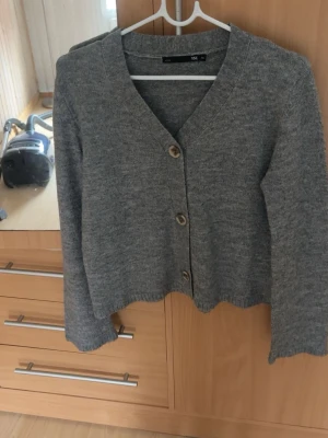 Grå kofta med knappar - Mysig grå kofta med v-ringning och tre stora träknappar framtill. Koftan har lång ärm och är stickad i mjukt material, perfekt för lager på lager. Enkel och stilren design som passar till det mesta.