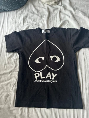 Svart Comme des Garçons PLAY t-shirt - Säljer en svart t-shirt från Comme des Garçons PLAY med det ikoniska hjärtat med ögon tryckt i vitt framtill. Klassisk passform och rund hals. Perfekt statement-plagg för dig som gillar streetwear och vill sticka ut.