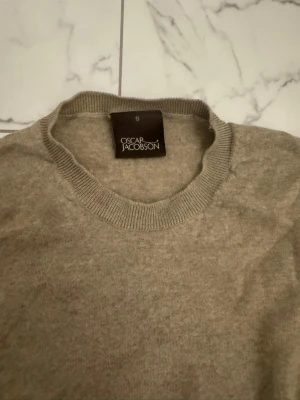 Beige stickad tröja Oscar Jacobson - Klassisk beige stickad tröja från Oscar Jacobson med rund halsringning och ribbade muddar vid ärmslut och nederkant. Perfekt för lager på lager och enkel att matcha med olika outfits. Mjuk och skön kvalitet.
