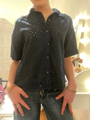 Mörkblå kortärmad skjorta med spets - Snygg mörkblå skjorta med korta ärmar och genombruten spetsdetalj. Skjortan har knappar framtill. Perfekt till sommarn med ett par shorts