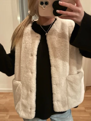 Krämvit fuskpälsväst med fickor - Säljer en krämvit pälsväst i mjuk fuskpäls med raka paneler och stora fickor framtill. Västen har en enkel, öppen design utan synliga knappar eller dragkedja. Perfekt att slänga över en hoodie för en chill och trendig look.