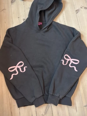 Svart hoodie med rosa detaljer Edikted - Cool svart hoodie från Edikted med huva och ribbade muddar. På båda ärmarna finns rosa tryck i form av rosetter som ger en unik touch. Tillverkad i mjukt material som känns skönt mot huden. Perfekt för dig som vill ha en stilren men ändå lekfull look.