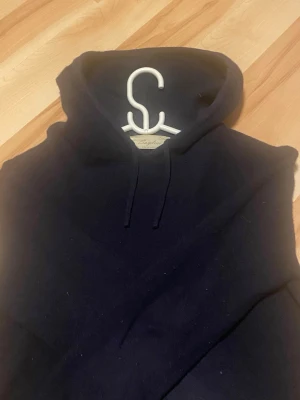 Mörkblå hoodie från Sayles - Snygg mörkblå hoodie från Sayles med klassisk huva och dragsko. Hoodien har en enkel och stilren design, perfekt för dig som gillar en clean look. Tillverkad i mjukt material som känns skönt mot huden. Passar perfekt till jeans eller joggers.
