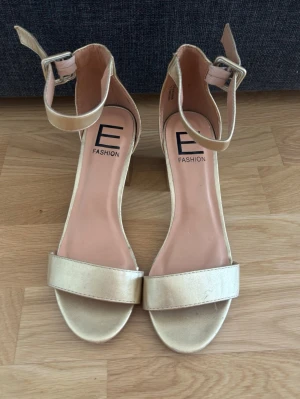 Guldiga blockklack - Snygga guldiga klackar frånmed bred rem över tårna och justerbart ankelband med spänne. 