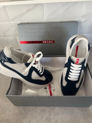 Prada sneakers  - Snygga sneakers från Prada i marinblått och vitt med en mix av glansigt läder och mesh-tyg. Klassisk låg modell med vita skosnören och robust sula. Ikonisk röd Prada-detalj på plösen och insidan. Perfekta för dig som gillar exklusiv streetstyle.  Pris kan ev disskuteras, enbart seriösa köpare. Kan vid efterfrågan även mötas i Stockholm. Allt är i nyskick