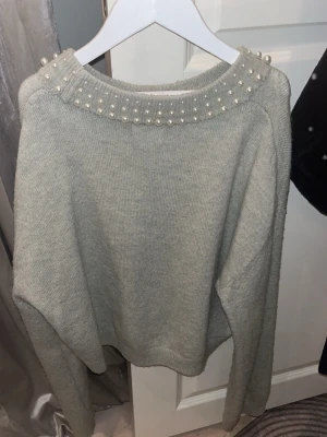 Grå cropped stickad tröja med pärlor - Snygg grå stickad tröja med cropped passform och pärldetaljer runt den breda halsringningen. Tröjan har långa ärmar och är gjord i mjukt material som känns skönt mot huden. Perfekt för dig som vill ha en trendig och bekväm look.