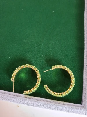 Guldiga creoler med gröna stenar - Stora creolörhängen i guldton med gnistrande gröna stenar runt hela ringen. Örhängena har en chunky design och ger ett riktigt statement-intryck. Perfekta för dig som vill sticka ut och addera färg till din look.