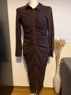 Brun långärmad skjortklänning - Mörkbrun långklänning med långa ärmar och krage. Klänningen har knappar hela vägen framtill, rynkade detaljer över bröstet och en slits nedtill. Materialet känns mjukt och stretchigt, troligen bomull eller polyester. Perfekt för dig som gillar en stilren och trendig look.