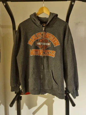 Grå Harley-Davidson zip hoodie New L - Mörkgrå hoodie med dragkedja från Harley-Davidson. Stor logga i orange och vitt på bröstet med texten 'Harley-Davidson Motorcycles'. Utrustad med huva och känguruficka framtill. Tillverkad i mjukt sweatshirtmaterial, perfekt för en avslappnad look.