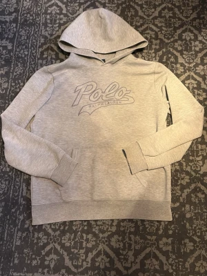 Ralph Lauren hoddie - Snygg ljusgrå hoodie från Polo Ralph Lauren med broderad logga framtill. Klassisk modell med huva och känguruficka. Perfekt för en avslappnad och stilren look. Mjuk bomullsblandning som är skön att ha på sig hela dagen. Storleken är 18-20 år vilket motsvarar S