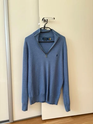 Blå halvzip-tröja från Polo Ralph Lauren - Snygg blå stickad tröja från Polo Ralph Lauren med halv dragkedja och broderad logga på bröstet. Tröjan har ribbade muddar och krage, perfekt för lager-på-lager-stil. Tillverkad i mjuk bomull för en bekväm känsla.