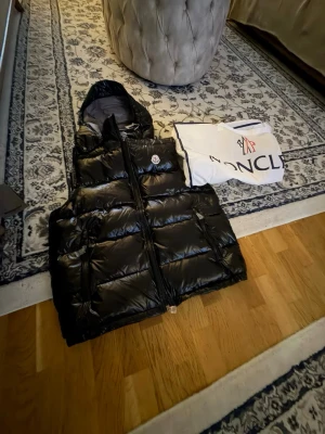Svart dunväst från Moncler med huva - Snygg svart dunväst från Moncler med glansig finish och avtagbar huva. Västen har två stora fickor framtill och dragkedja. Klassisk Moncler-logga på bröstet och medföljande dustbag. Perfekt för lager-på-lager-stil under kyliga dagar.