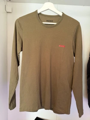 Beige långärmad topp från Hugo - Snygg beige långärmad topp från Hugo med rund halsringning och diskret röd logga på bröstet. Tillverkad i mjuk bomull som känns skön mot huden. Perfekt för dig som gillar stilrena och enkla plagg med en touch av branding.