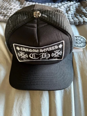 Svart Chrome Hearts keps med mesh - Snygg svart keps från Chrome Hearts med vit brodyr framtill och på baksidan. Kepsen har meshpaneler och justerbar snapback. Metallblomma på toppen ger extra edge. Perfekt för dig som gillar streetwear och vill sticka ut.