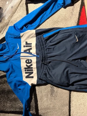 Nike Air blå och mörkblå träningsset M-L - Nike Air träningsset med hoodie och byxor i blått och mörkblått. Hoodien har dragkedja, huva och stor Nike Air-logga framtill. Byxorna har resår i midjan och dragsko. Perfekt för gymmet eller chill. Materialet är mjukt och stretchigt för maximal komfort.