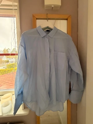 Ljusblå skjorta med bröstficka - Klassisk ljusblå skjorta med krage och dold knappslå. Skjortan har lång ärm och en bröstficka på vänster sida. Perfekt för en stilren och enkel look.