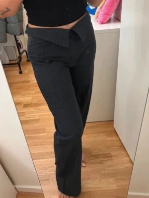 Svarta kostymbyxor med vit rand - Säljer ett par svarta kostymbyxor med vita tunna kritstreck och låg midja. Byxorna har vida ben och en unik asymmetrisk linning framtill. Perfekta för dig som vill ha en trendig och stilren look.