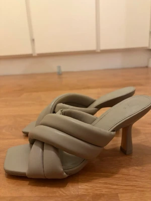 Beige quiltade pumps med klack - Snygga pumps i beige med trendig quiltad design och breda korslagda remmar. Skorna har fyrkantig tå och medelhög klack, vilket ger en modern och stilren look. Tillverkade i ett mjukt material som liknar läder. Små i storlek