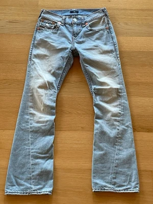 True Religion Jeans - Säljer ett par ljusblå jeans från True Religion i modellen Joey Flare. Jeansen har låg midja, snygga detaljer på bakfickorna och är tillverkade i bomull med en lätt tvättad look. De har pyttelite slitage ned till vid benöppningen, men bortsett från det mycket fint skick. Längden på byxorna är L32.