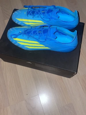 Adidas f50 elite Ag - Säljer ett par snygga blåa Adidas fotbollsskor med gula ränder och detaljer. Skorna har snörning och mönstrad ovansida i syntetmaterial samt fasta dobbar för spel på gräs. Perfekta för dig som vill sticka ut på planen!