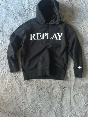 Svart hoodie från Replay - Svart hoodie från Replay med stor vit logga på bröstet. Klassisk modell med huva och dragsnören, samt ribbade muddar vid ärmslut och nederkant. Perfekt för en avslappnad och trendig stil.