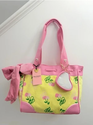 Juicy Couture daydreamer tote rosa och gul  - Superfin handväska från Juicy Couture i rosa och gult med körsbärsmönster och gröna detaljer. Väskan har rosa handtag, en stor rosett i tyg och ett hjärtformat spegelhänge. Materialet är en mix av tyg och konstläder.
