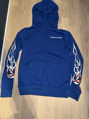 Chrome heart - Snygg blå hoodie från Crone Worldz med huva och känguruficka. Ärmarna har coola tryck med blåa och röda munnar, och på ryggen finns ett stort motiv med texten 'SPACE' och en tecknad figur. Perfekt för dig som gillar streetwear och unika prints. STORLEK S MEN PASSAR SPM XS!!!