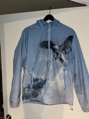 Blå vindjacka med örntryck från Trapstar - Snygg ljusblå vindjacka från Trapstar med stort örntryck på både fram- och baksida. Jackan har huva, dragkedja framtill och två fickor med dragkedja. På ryggen står det 'TRAPSTAR' i stora bokstäver. Perfekt för en streetwear-look.