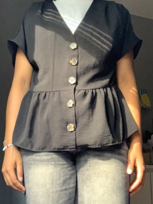 Svart blus med knappar och v-ringning - Svart blus med v-ringning och stora dekorativa knappar framtill. Modellen har korta ärmar och en volang nedtill som ger en snygg siluett. Perfekt att styla med jeans eller kjol för en trendig look.
