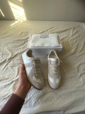 Maison Margiela vita sneakers - Säljer ett par stilrena Maison Margiela sneakers i vitt och beige med klassisk gummisula. Skorna har en mix av slätt läder och mocka, samt vita snören. Perfekta för dig som vill skapa en så clean och minimalistisk outfit som möjligt. Skick 9.5/10 endast använda inomhus. Box + dustbag följer med. Säljer på grund av fel storlek