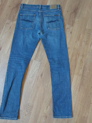 Nudie jeans lean dean - Ett par klassiska blå jeans från Nudie Jeans med raka ben och femficksmodell. Jeansen har tydliga sömmar, läderpatch bak i midjan och signaturbroderi på bakfickorna. Tillverkade i slitstark denim, perfekta för en avslappnad stil.