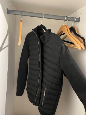 Svart pufferjacka från Moncler - Snygg svart pufferjacka från Moncler med quiltad framsida, slät baksida och ärmar. Jackan har dragkedja framtill, hög krage och en ficka med dragkedja. Moncler-logga på ärmen ger en exklusiv touch. Perfekt för kalla vinterdagar.