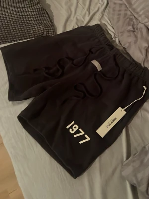 Svarta Essentials shorts med snörning - Svarta shorts från Essentials med vit '1977'-print på ena benet och snörning i midjan. De har en relaxed passform och är tillverkade i mjukt material som passar perfekt för chill eller träning. Elastisk midja för extra komfort.
